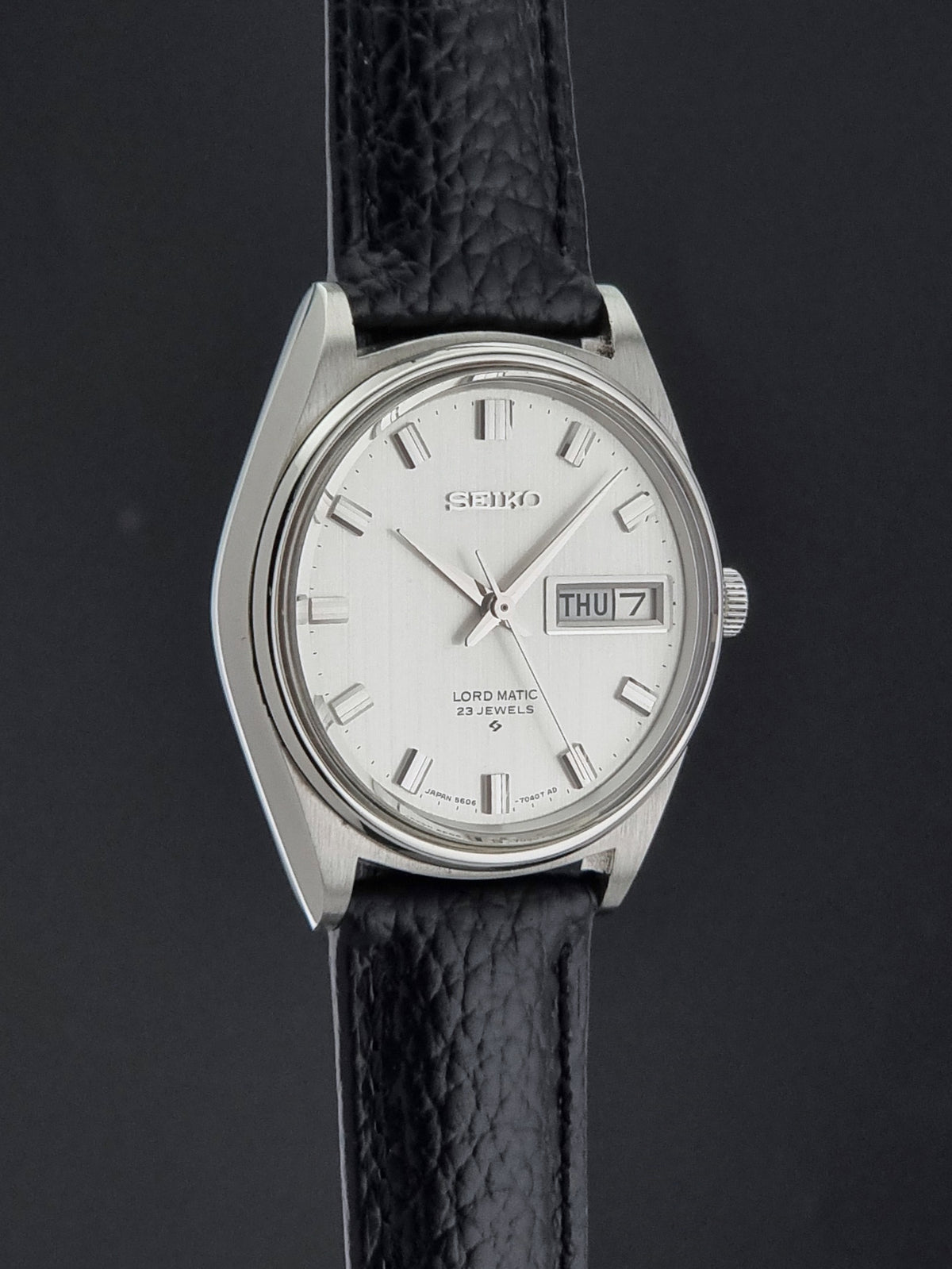 A Vintage Seiko watch LordMatic 5606-7040 with a silver-tone case, white dial, and black textured leather strap, shown against a dark gray background.