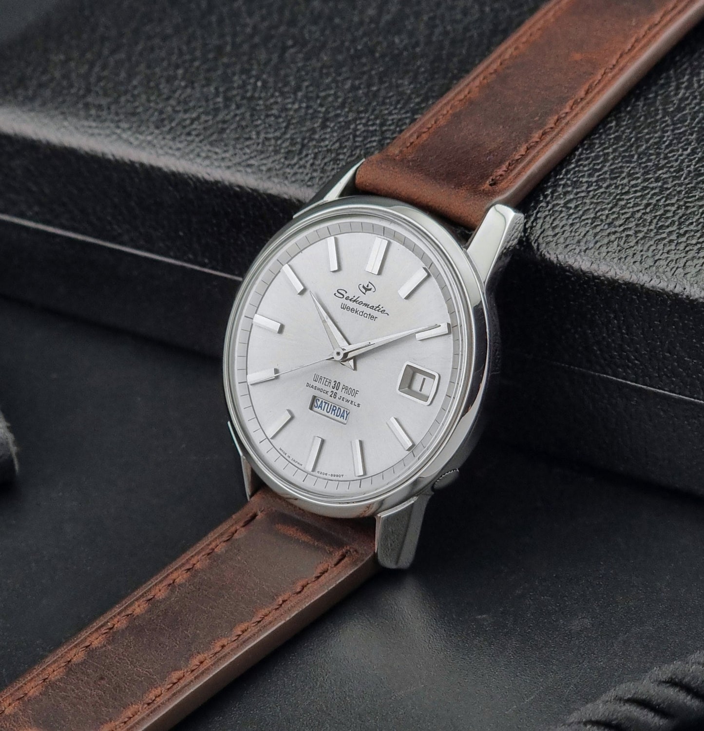 Vintage Seiko watch Seikomatic Weekdater 6206-8990 with brown leather strap on a gray textured background crown view