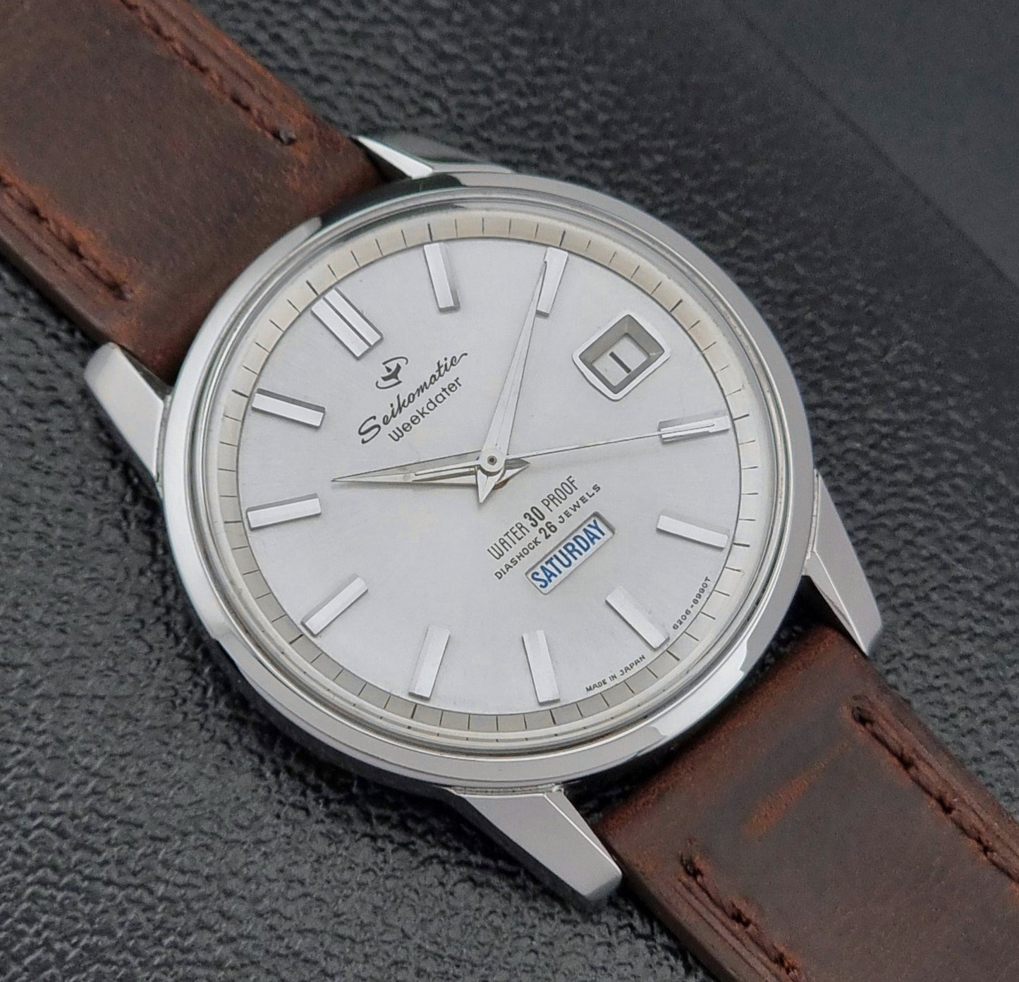 Vintage Seiko watch Seikomatic Weekdater 6206-8990 with brown leather strap on a gray textured background dial view
