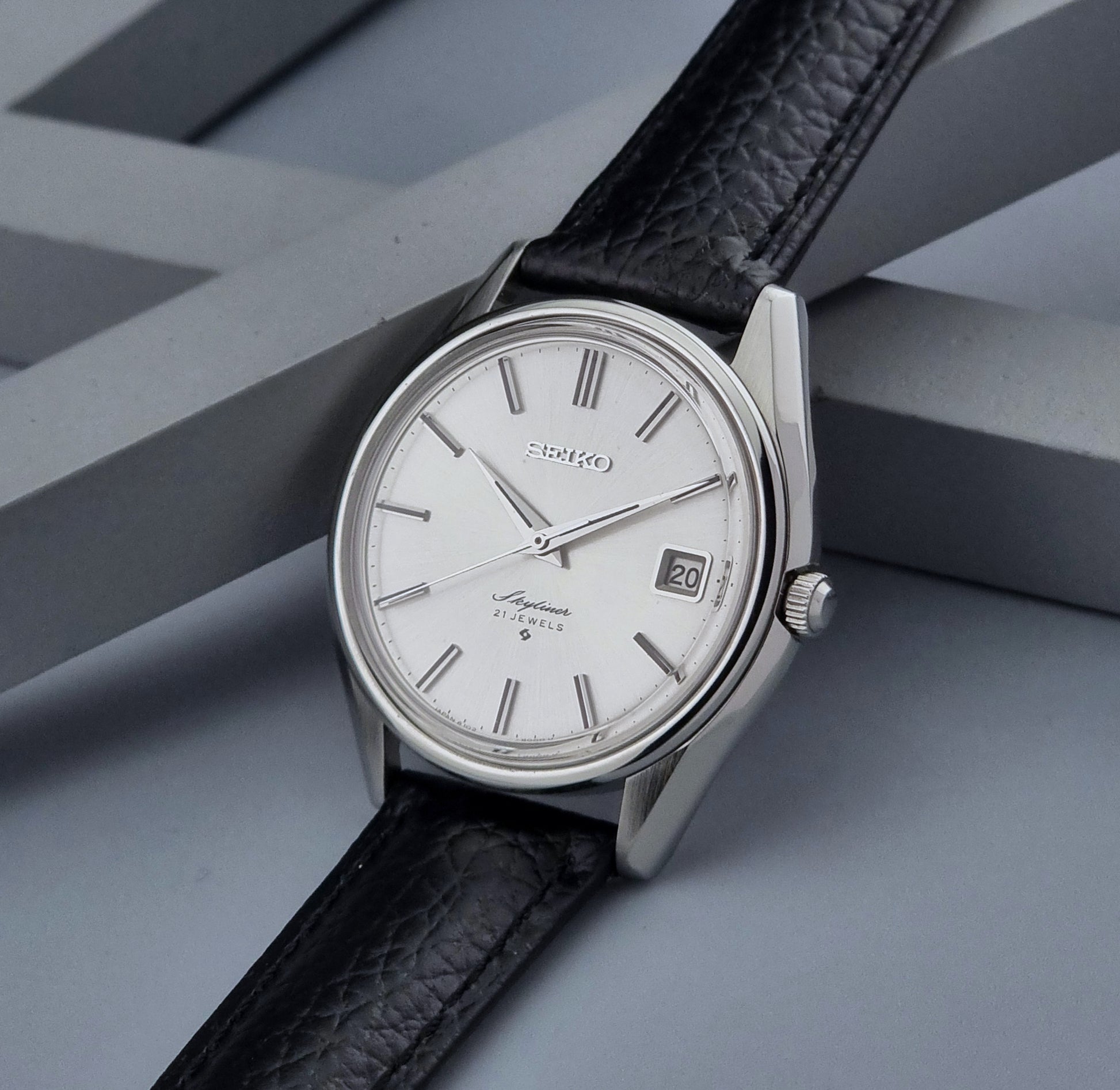 Vintage Seiko Wristwatch Skyliner 6102-8000 with black leather strap and silver case on a light gray background crown view