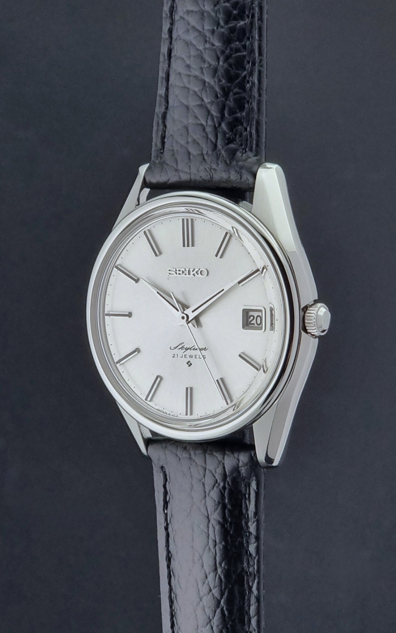 Vintage Seiko Wristwatch Skyliner 6102-8000 with black leather strap and silver case on a light gray background crown profile view