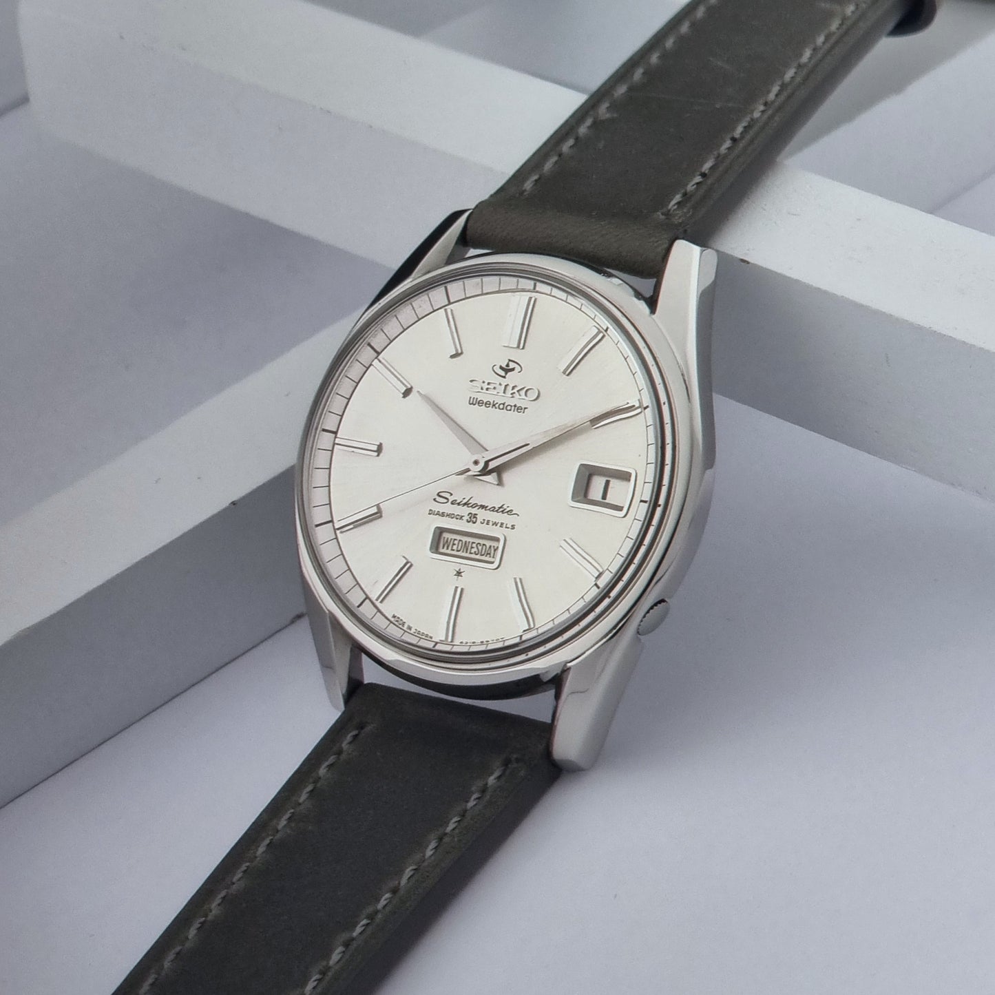 Vintage Seiko watch Seikomatic Weekdater 6218-8970 with a silver case and dark leather strap on a light gray background crown view