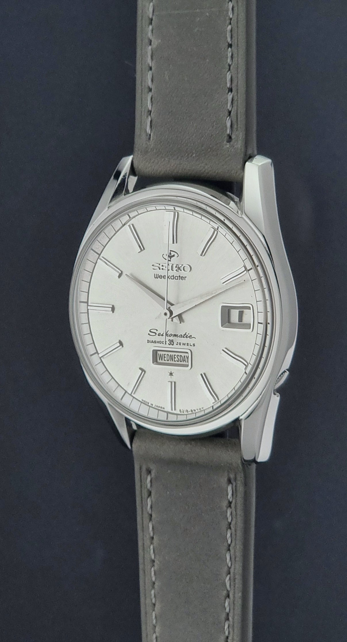 Vintage Seiko watch Seikomatic Weekdater 6218-8970 with a silver case and dark leather strap on a light gray background crown profile view