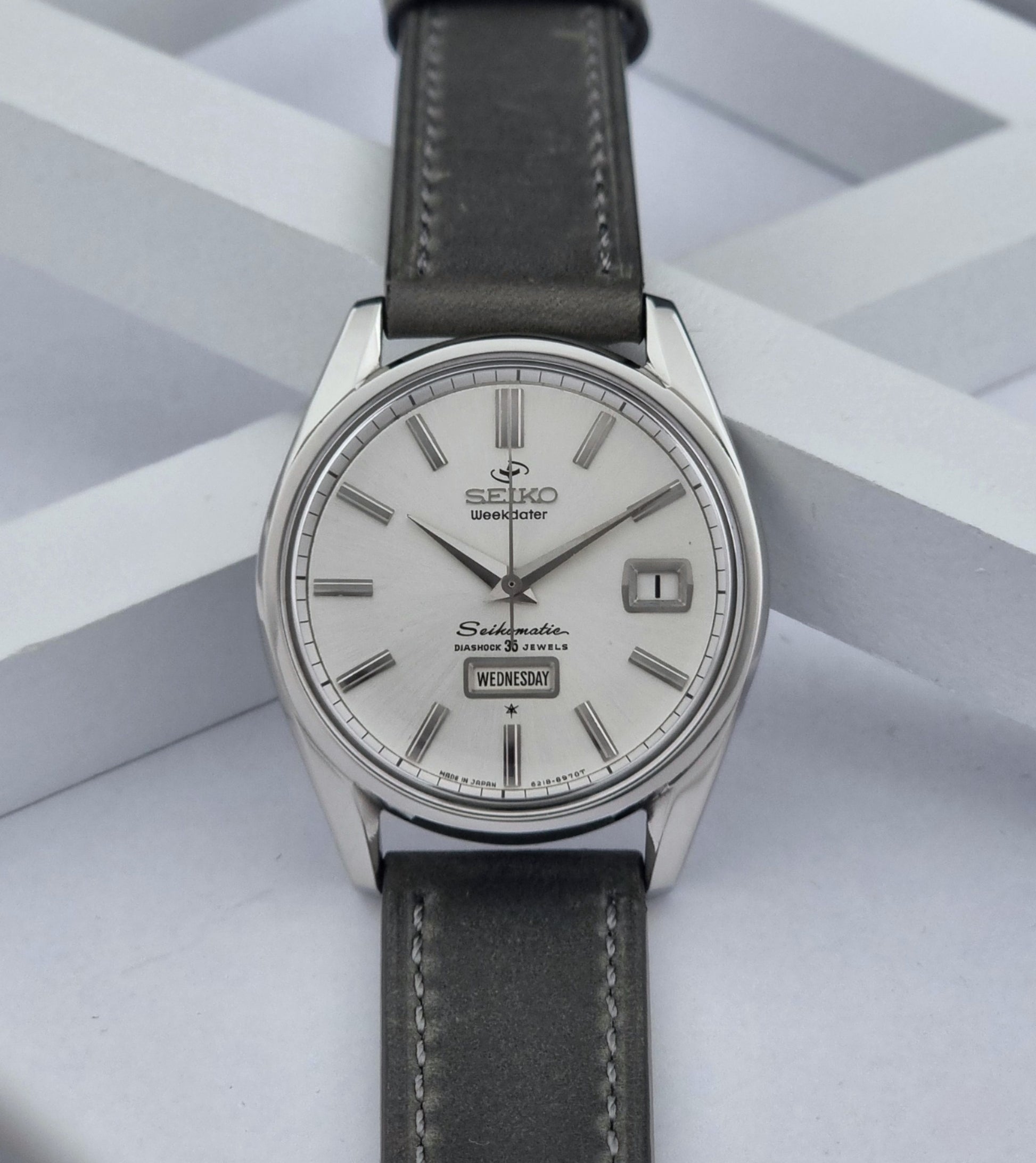 Vintage Seiko watch Seikomatic Weekdater 6218-8970 with a silver case and dark leather strap on a light gray background dial view