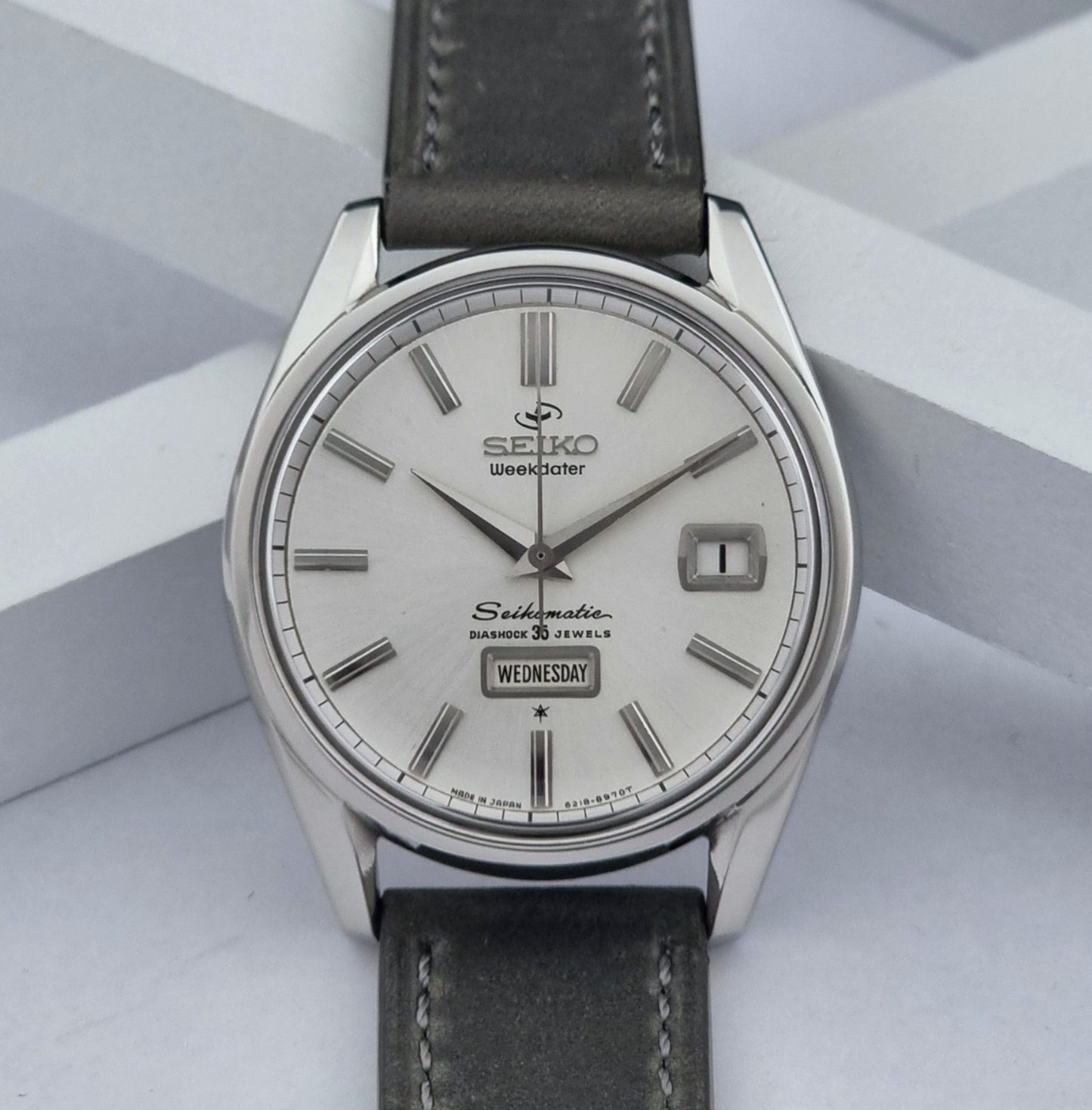 Vintage Seiko watch Seikomatic Weekdater 6218-8970 with a silver case and dark leather strap on a light gray background front view