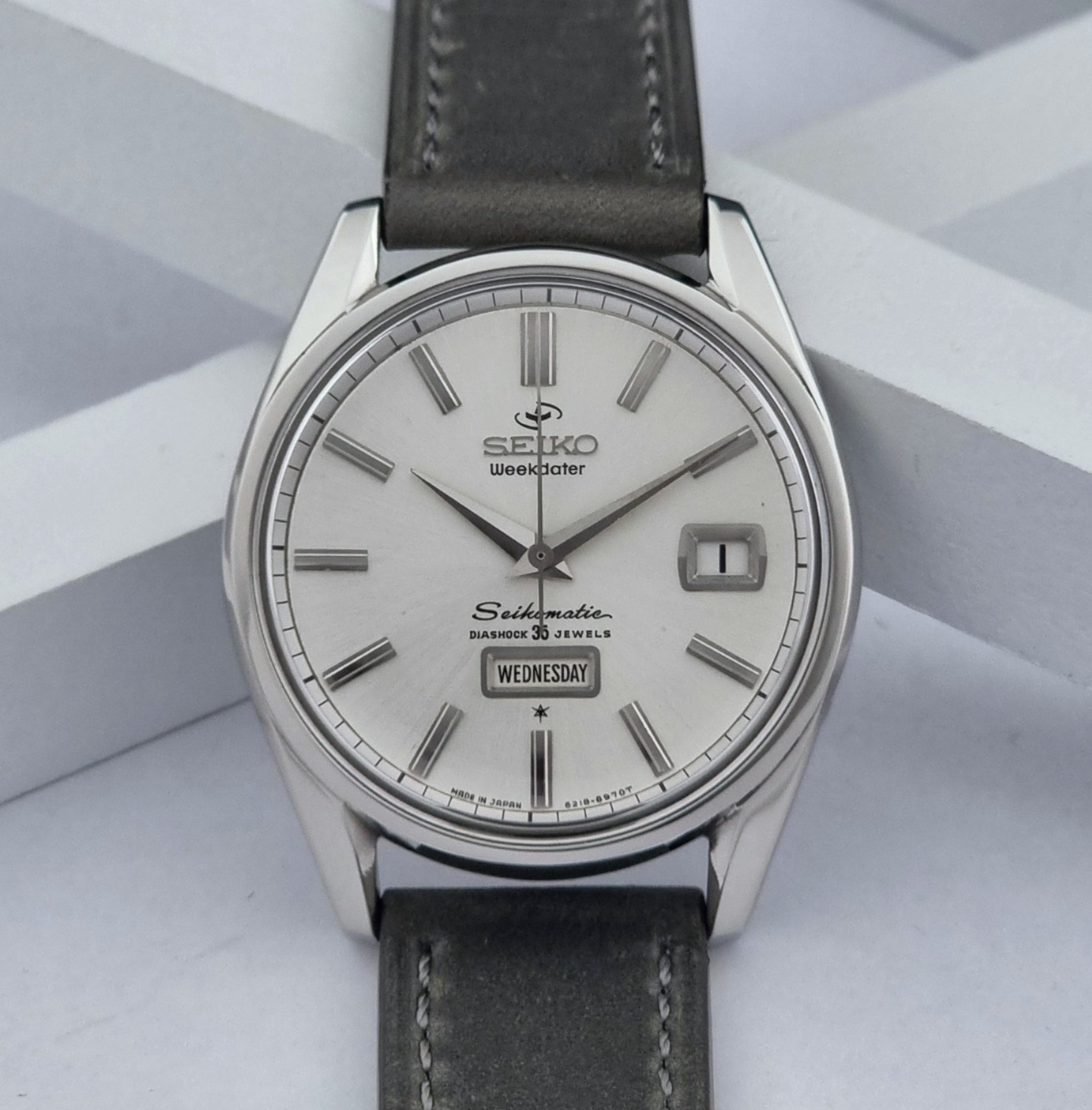 Vintage Seiko watch Seikomatic Weekdater 6218-8970 with a silver case and dark leather strap on a light gray background front view