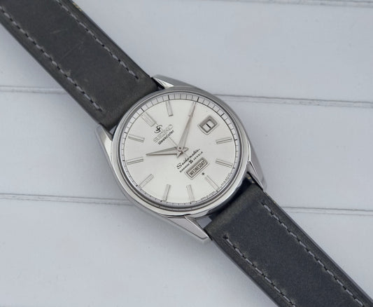 Vintage Seiko watch Seikomatic Weekdater 6218-8970 with a silver case and dark leather strap on a light gray background dial