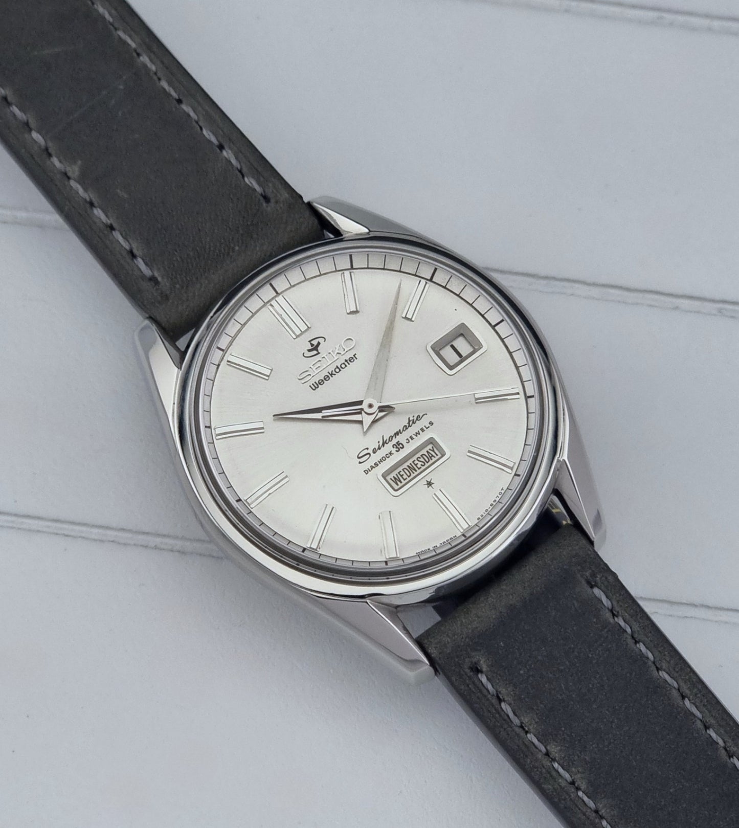 Vintage Seiko watch Seikomatic Weekdater 6218-8970 with a silver case and dark leather strap on a light gray background close up dial view