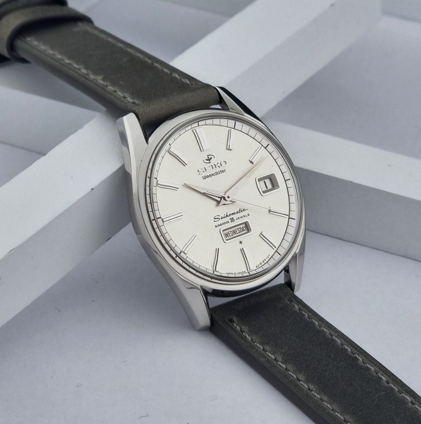 Vintage Seiko watch Seikomatic Weekdater 6218-8970 with a silver case and dark leather strap on a light gray background side view