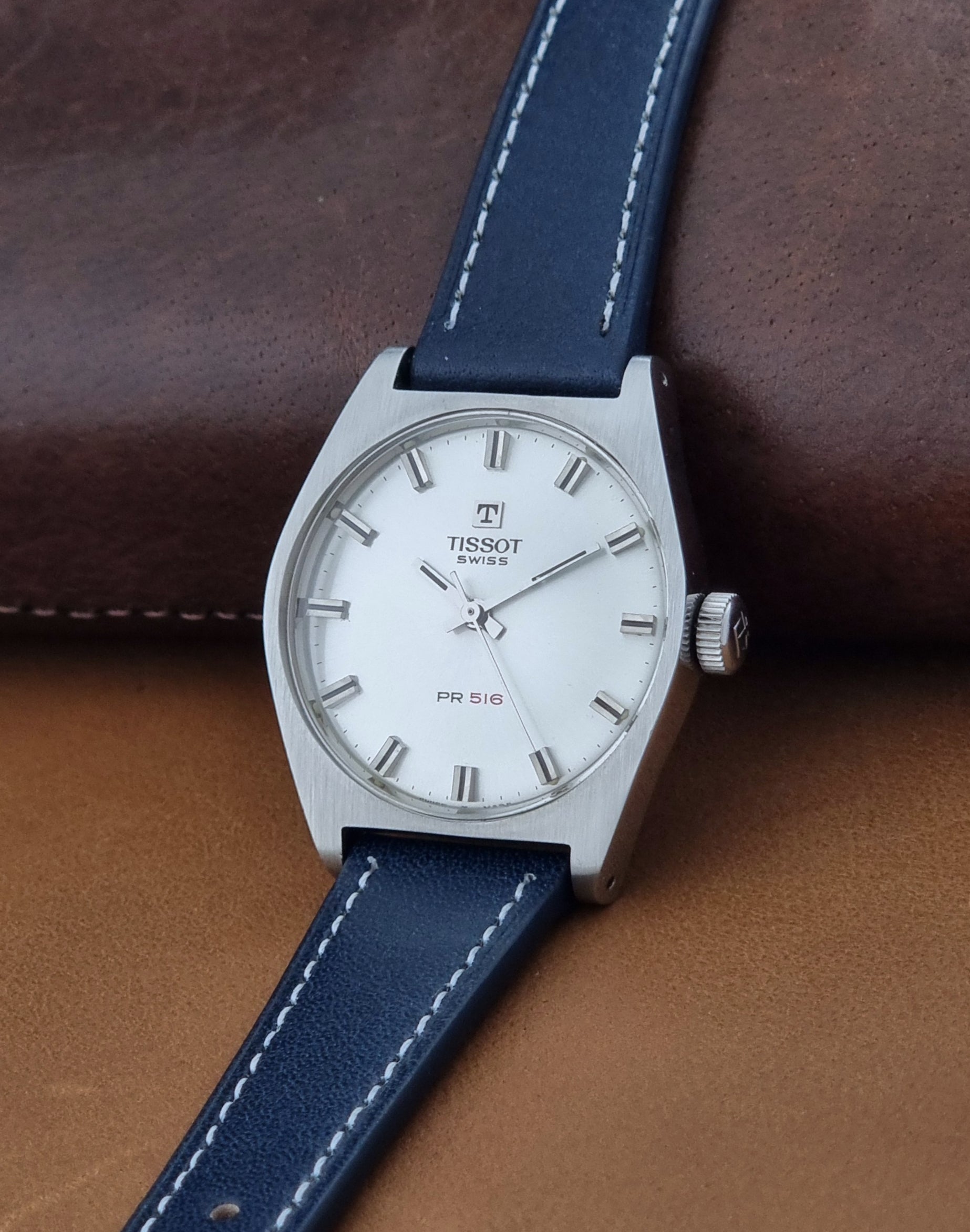 A Tissot Swiss PR 516 watch with a silver-tone case, white dial, and a dark blue leather strap with white stitching, resting on a brown leather surface.