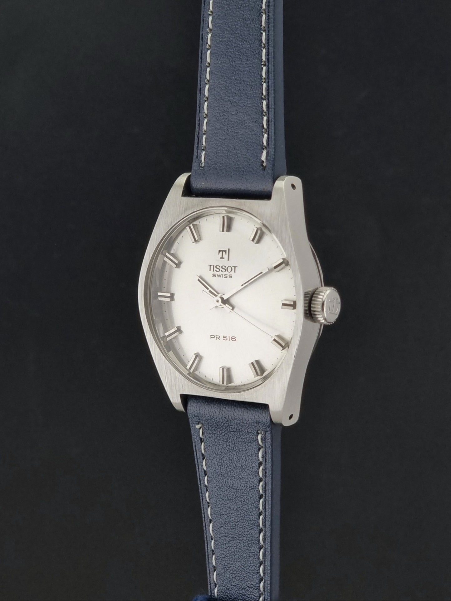 Vintage Tissot Wristwatch 41620 PR 516 watch with a silver-tone metal case, white dial, and a blue leather strap with white contrast stitching, shown against a dark background.