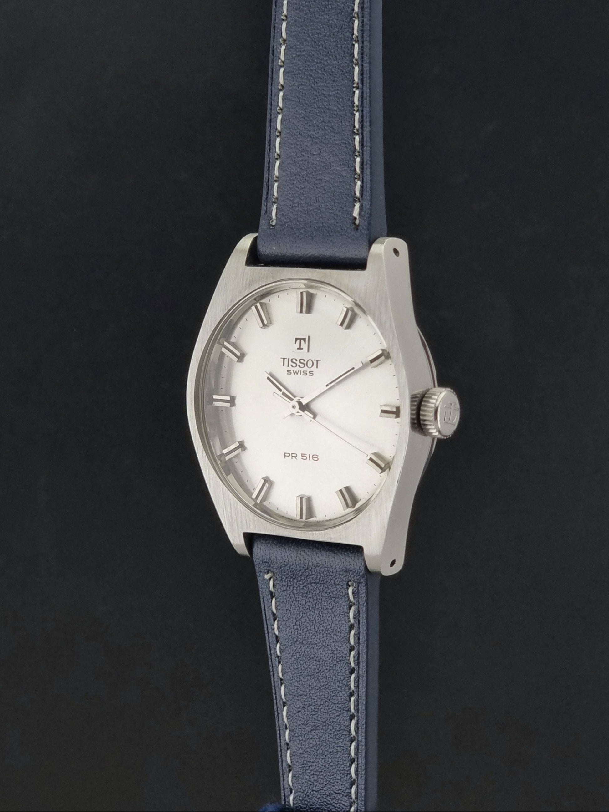 Vintage Tissot Wristwatch 41620 PR 516 watch with a silver-tone metal case, white dial, and a blue leather strap with white contrast stitching, shown against a dark background.