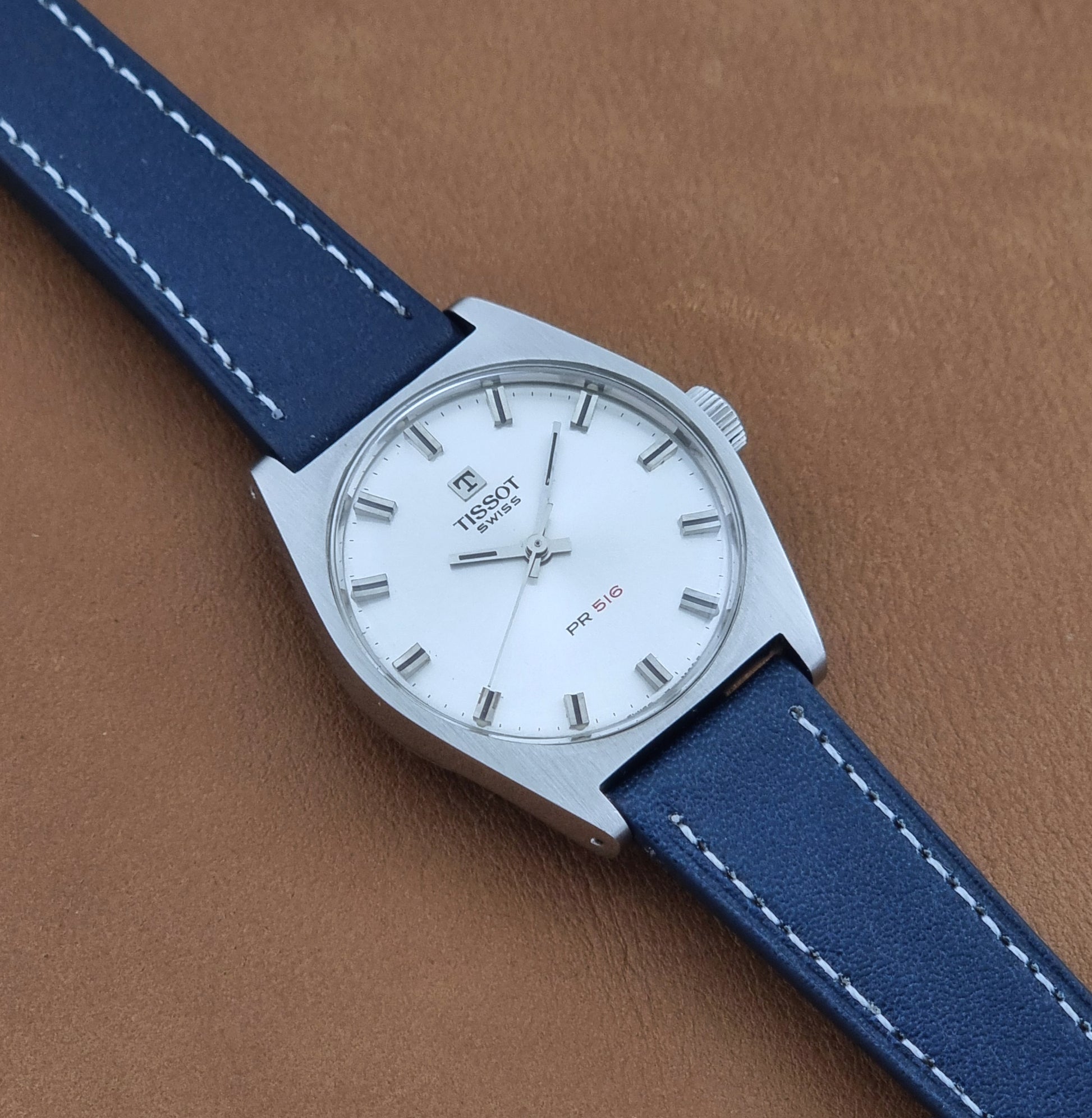 Vintage Tissot Wristwatch 41620 PR 516 watch with a silver-tone stainless steel case, white dial, and blue leather strap with white stitching, displayed diagonally on a brown textured leather background.