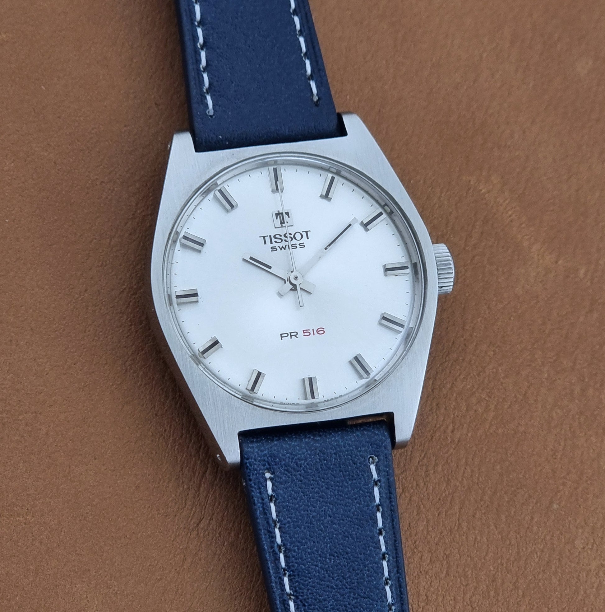 Vintage Tissot Wristwatch 41620 PR 516 watch with a silver stainless steel case, white dial, and a blue leather strap with white stitching, shown on a brown textured background.