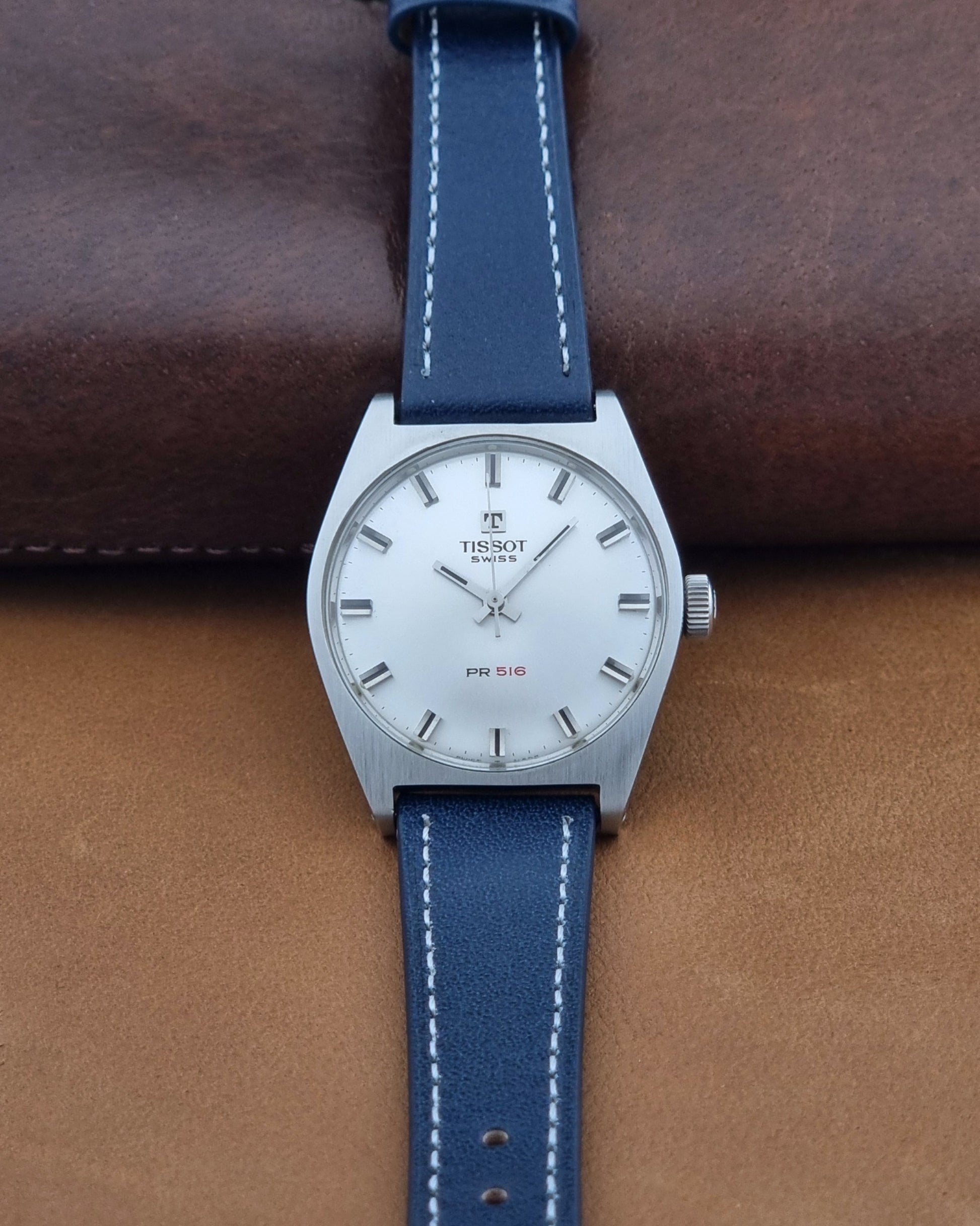 A Tissot Swiss PR 516 watch with a silver stainless steel case and a blue leather strap with white stitching, displayed on a brown leather surface.