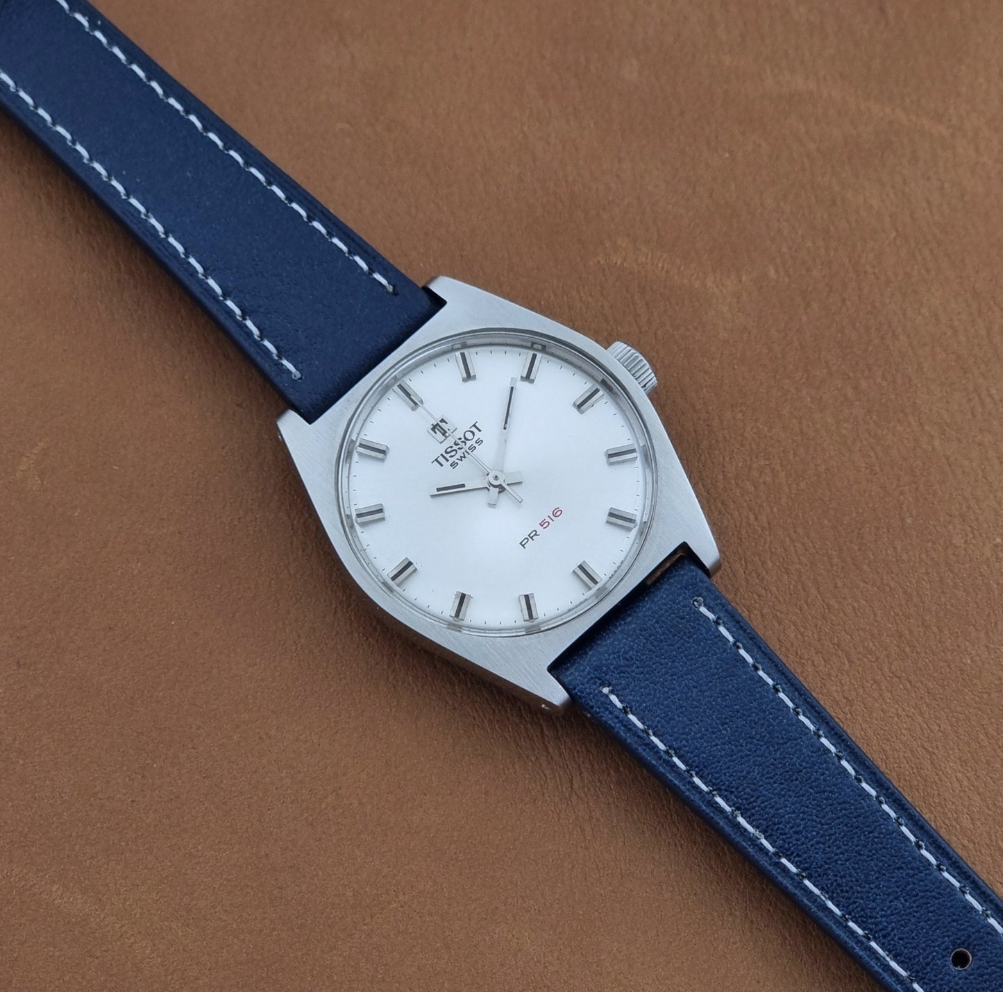 Vintage Tissot Wristwatch 41620 PR 516 watch with a silver-toned case, and blue leather strap with white stitching, displayed diagonally on a brown textured leather background.