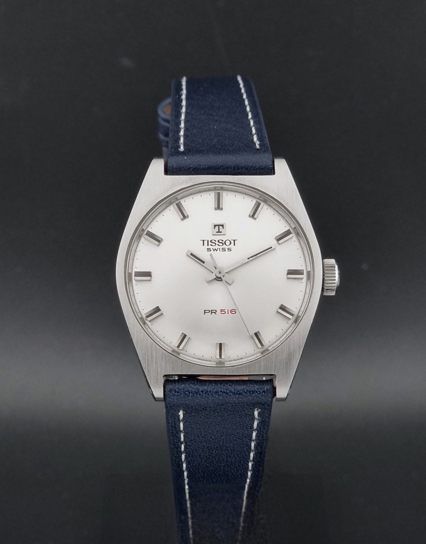 A vintage Tissot PR 516 wristwatch with a silver-tone metal case, white dial, and a dark blue leather strap with white stitching, shown against a dark gray background.