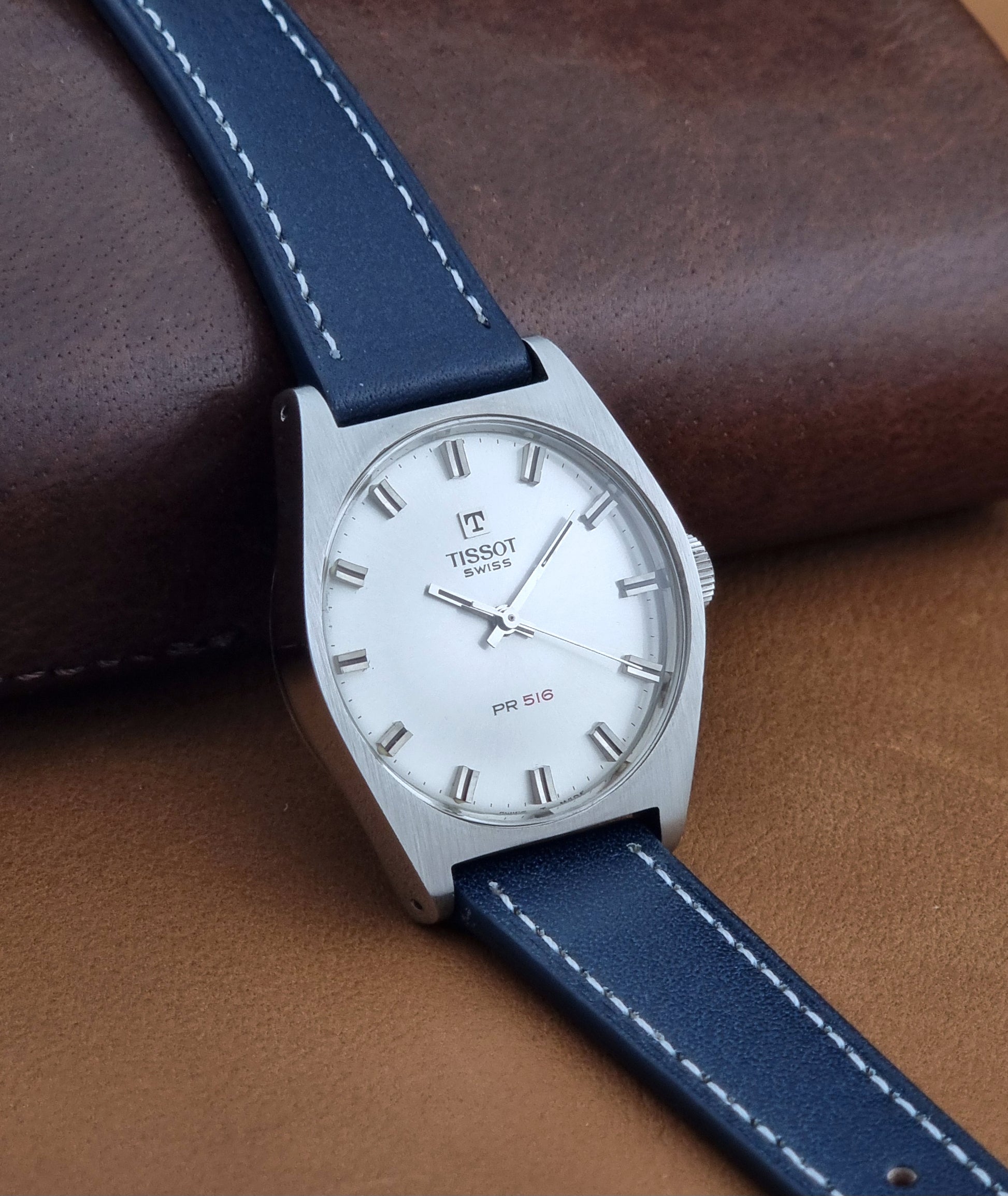 Vintage Tissot Wristwatch 41620 PR 516 watch with a silver-tone case, white dial, and dark blue leather strap with white stitching, resting on a brown leather surface.