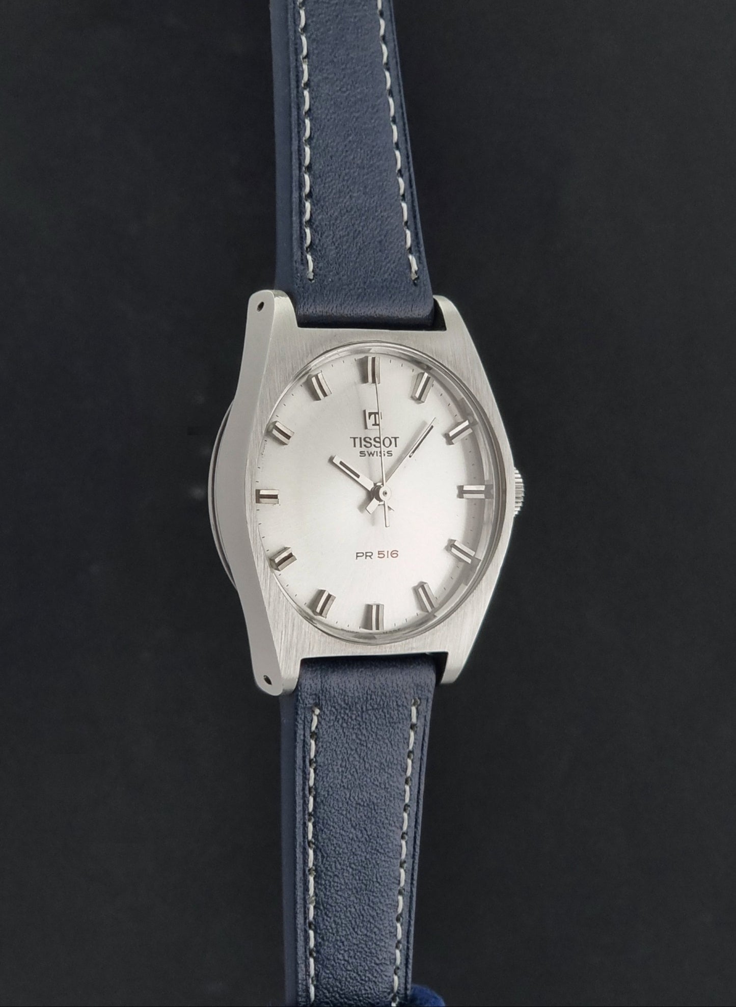 Vintage Tissot Wristwatch 41620 PR 516 wristwatch with a silver-tone oval case, white dial, and a blue leather strap with white stitching, shown against a dark background.