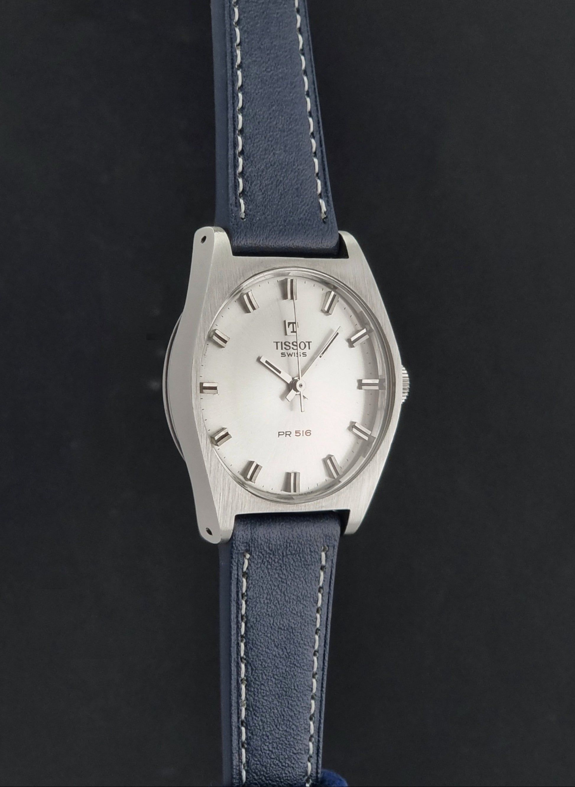 Vintage Tissot Wristwatch 41620 PR 516 wristwatch with a silver-tone oval case, white dial, and a blue leather strap with white stitching, shown against a dark background.