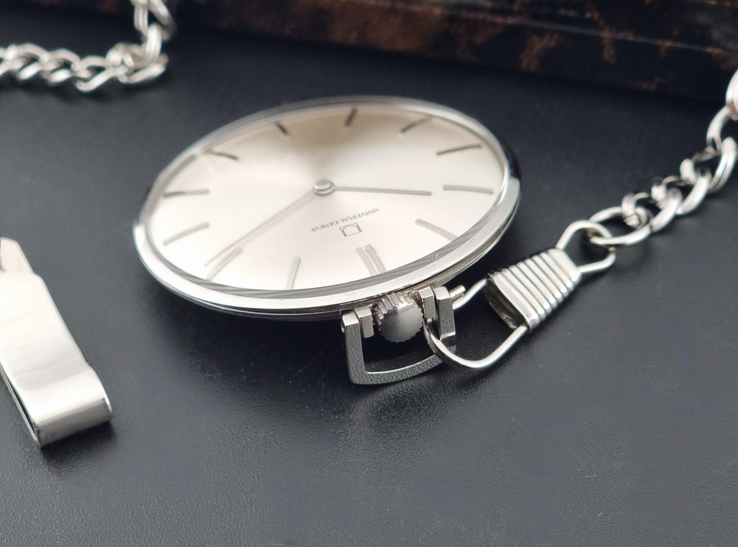 A Universal Geneve 856105/01 silver-toned pocket watch with a matching chain and a small metal accessory, photographed on a dark textured surface.