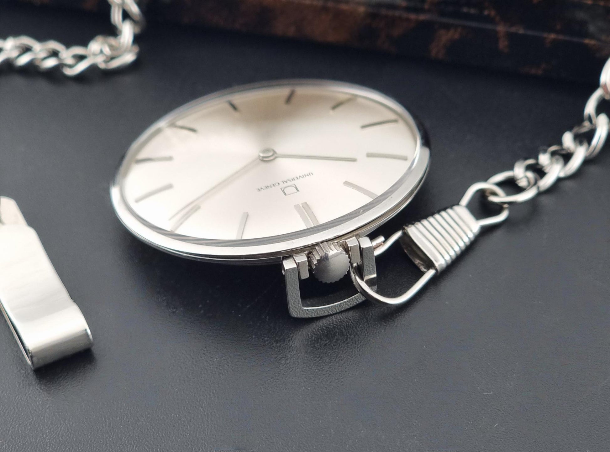 A Universal Geneve 856105/01 silver-toned pocket watch with a matching chain and a small metal accessory, photographed on a dark textured surface.