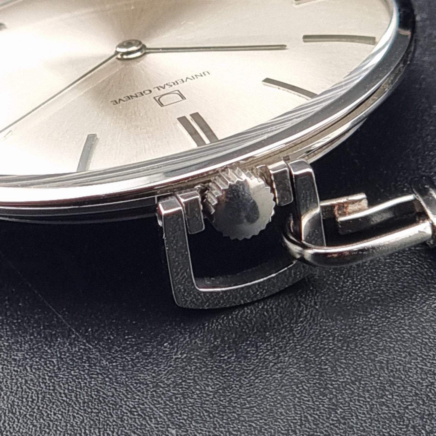 A Universal Geneve 856105/01 close-up of a silver-toned watch showing the dial, crown, and lugs against a dark textured surface. The dial has a brushed silver finish with simple rectangular hour markers and the text 'UNIVERSAL-GENEVE' visible.