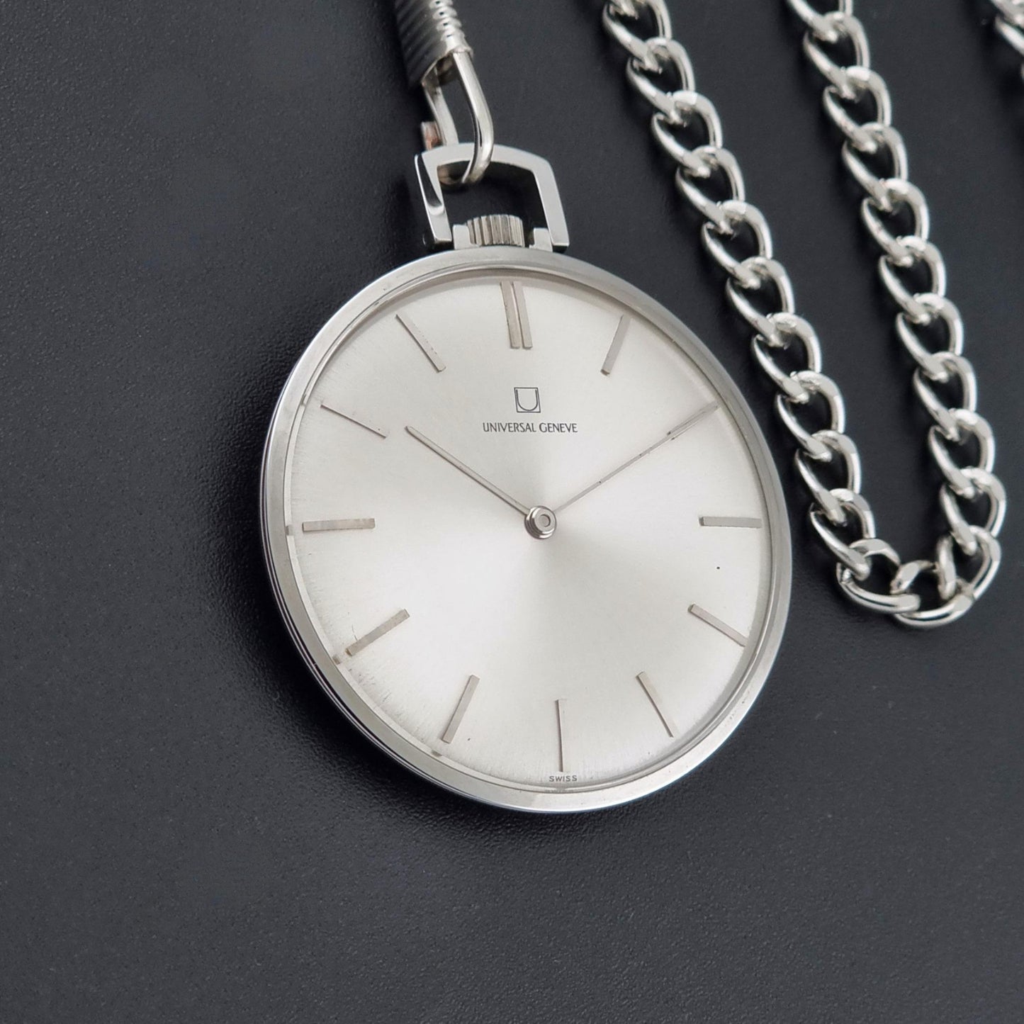 A silver analog pocket watch with a white dial and slim stick indices, shown with its matching silver chain on a dark textured background. The brand name Universal Geneve is visible on the watch face.