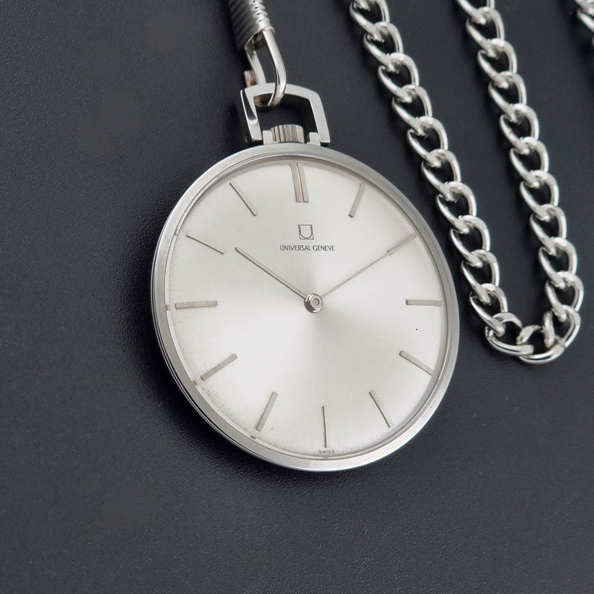 A silver analog pocket watch with a white dial and slim stick indices, shown with its matching silver chain on a dark textured background. The brand name Universal Geneve is visible on the watch face.