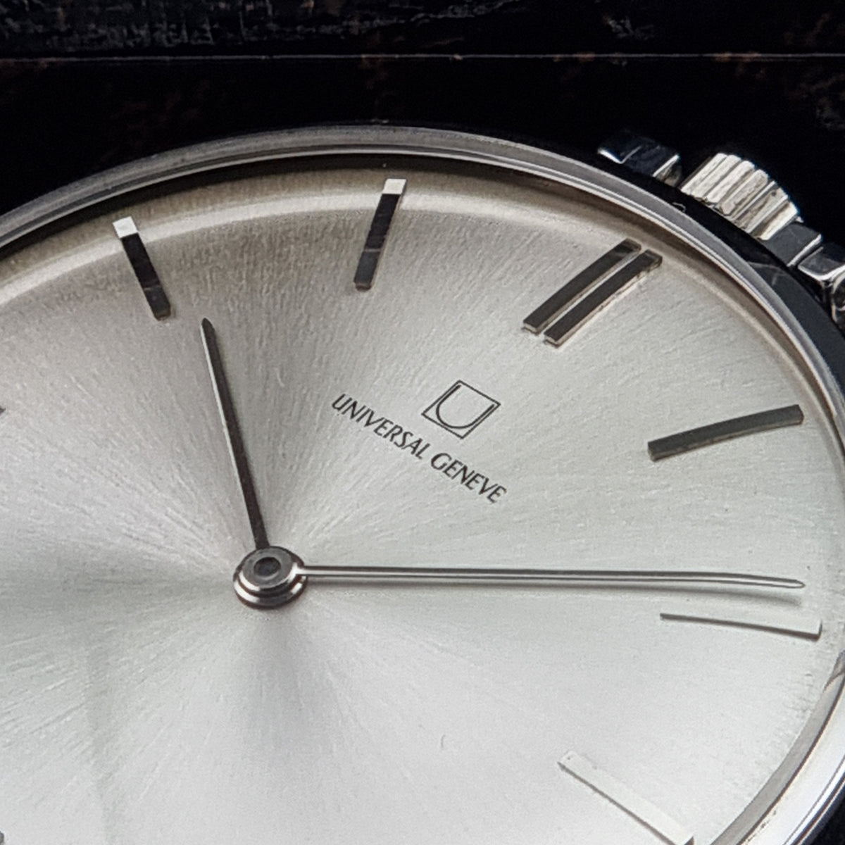 Close-up of a Universal Genève wristwatch dial with a brushed silver sunburst finish, slim baton hour markers, and silver-tone hands on a dark background.