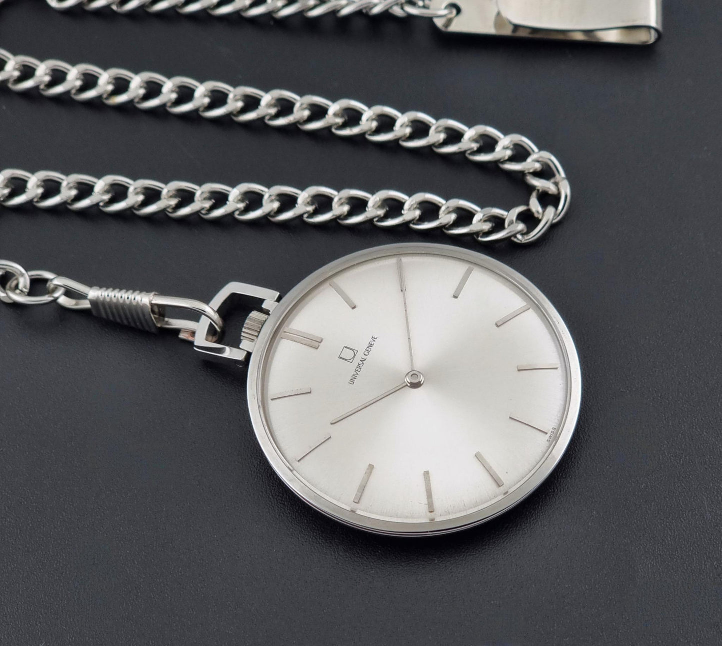 A Universal Geneve 856105/01  silver stainless steel pocket watch with a round silver dial and matching chain, displayed on a dark textured background.