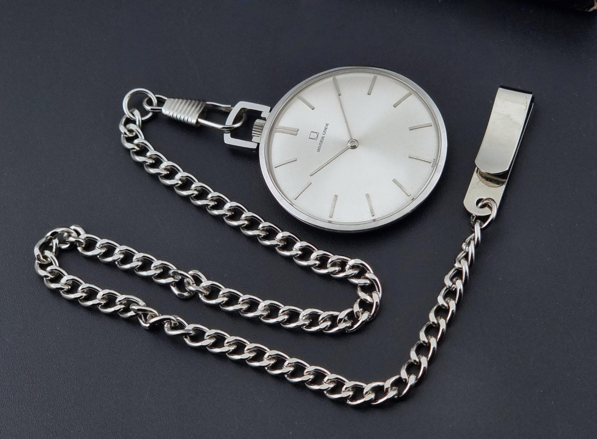 A Universal Geneve 856105/01 silver coloured pocket watch with a white dial and a matching silver chain, displayed on a dark gray background.