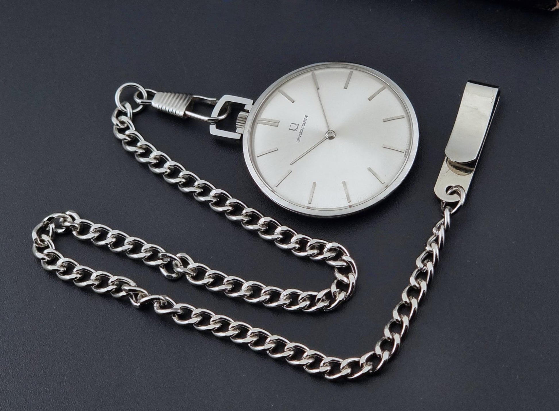 A Universal Geneve 856105/01 silver coloured pocket watch with a white dial and a matching silver chain, displayed on a dark gray background.